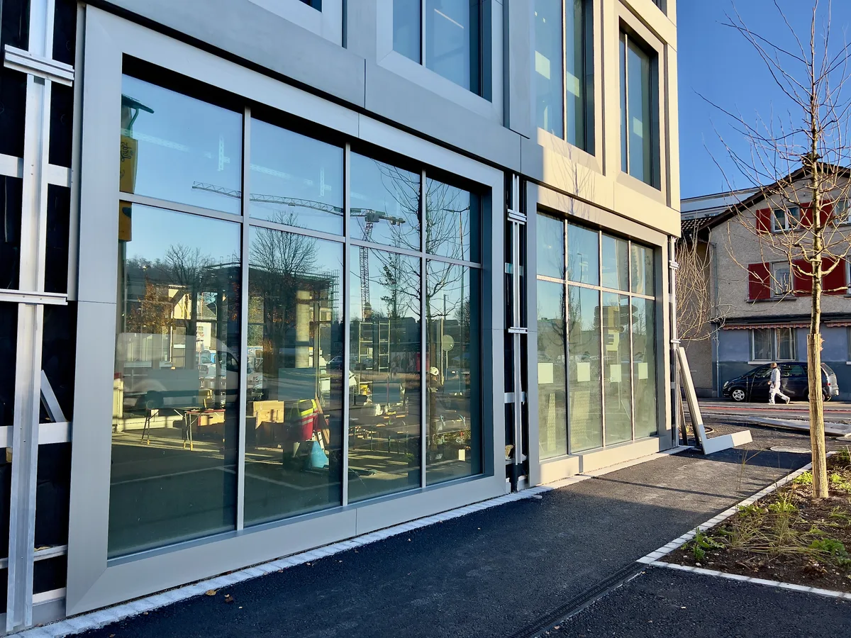 Man sieht die Fensterfront eines Neubaus neben einem Gehweg. Im Hintergrund ist ein Wohnhaus auf der gegenüberliegenden Strassenseite zu sehen.  